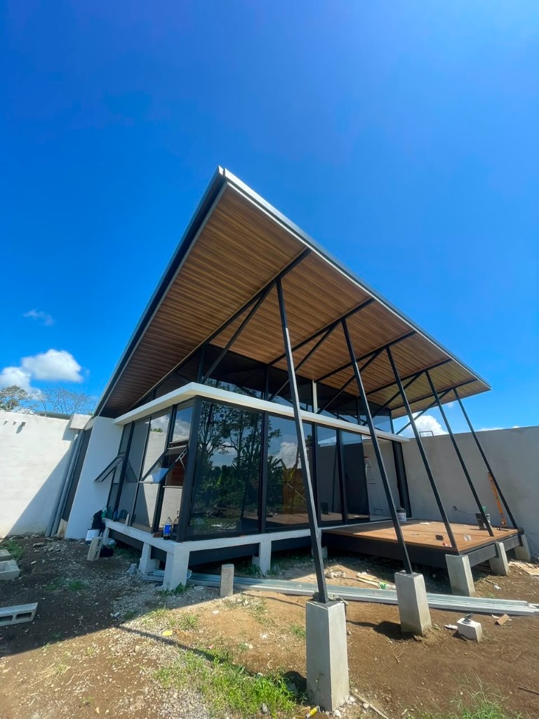 Construction progress — dramatic exterior view of the full building with sloped wood ceiling, steel framing, and glass walls against a blue sky